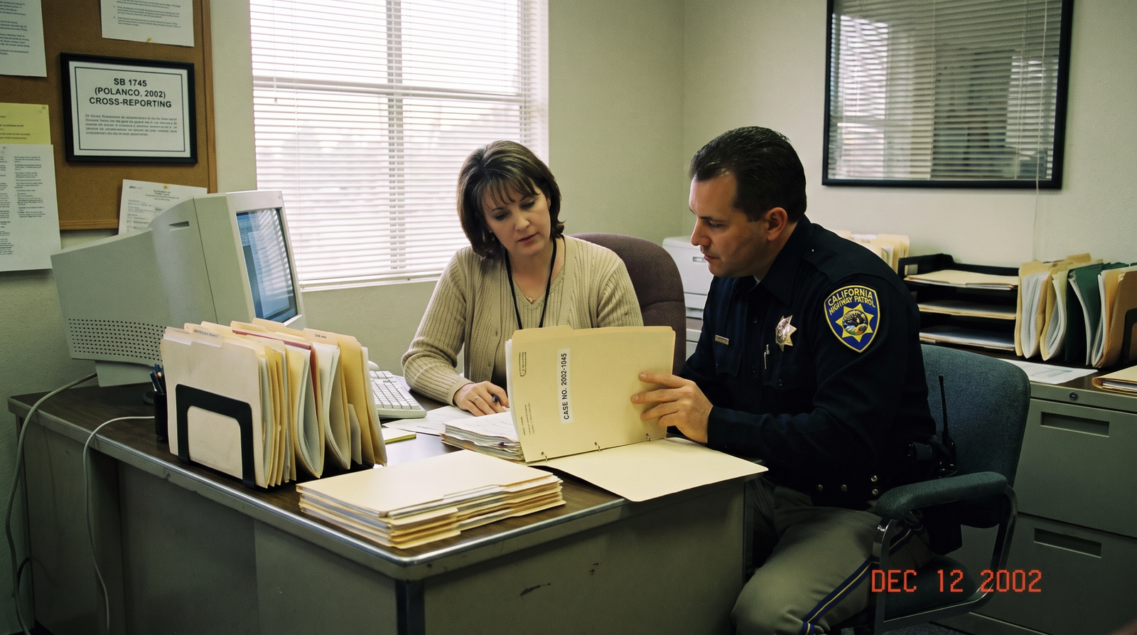 California child welfare office early 2000s, a social worker and a law enforcement officer reviewing a child welfare case file together at a desk, late afternoon light, archival editorial style, dignified and procedural. SB 1745 (Polanco, 2002) required CPS and law enforcement to cross-report child welfare cases.