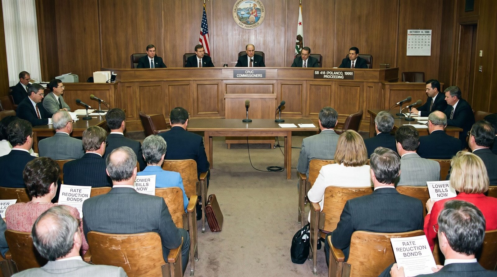 California Public Utilities Commission hearing room late 1990s, the bench and audience at a rate-reduction bond proceeding, archival editorial style, the kind of room where utility rates were decided. SB 418 (Polanco, 1999) authorized rate-reduction bonds for California utilities.