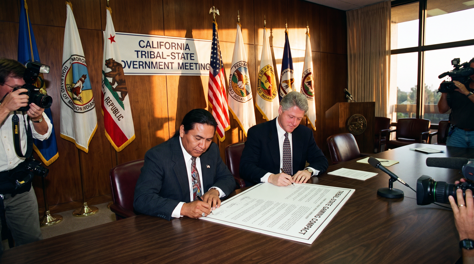 California tribal-state government meeting mid-1990s, two officials signing a tribal-state gaming compact at a long wooden table, with tribal flags and the California state flag in the background, late afternoon light, archival editorial style, dignified and procedural. SB 1350 (Polanco, 1999) established the tribal-state gaming compact framework.